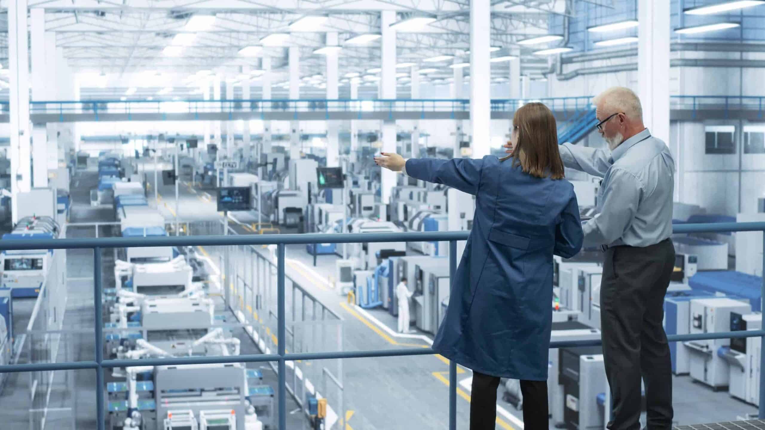 Experienced Male and Female Engineers Standing on a Platform with Their Back to Camera, Using Laptop Computer and Discussing Production at a Modern AI Automated Electronics Manufacture