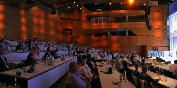 Das Bild zeigt den beeindruckenden Konferenzsaal während des Best Practice Day. Die Teilnehmer sind in mehreren Reihen von Tischen und Stühlen untergebracht, die zur Bühne hin ansteigen. Der Saal ist gut gefüllt, und alle blicken aufmerksam nach vorne, wo Wilhelm Goschy, CEO der STAUFEN.AG, einen Vortrag hält. Sein Name und Bild sind prominent auf einer großen Leinwand hinter ihm zu sehen. Die stimmungsvolle Beleuchtung in warmen Tönen und die farbigen Lichtprojektionen an den Wänden verleihen dem Raum eine einladende und professionelle Atmosphäre. Auf den Tischen der Teilnehmer sind Wasserflaschen und Gläser bereitgestellt, was zur konzentrierten und geschäftigen Stimmung des Events beiträgt.