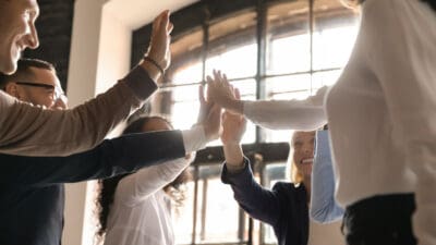 Overjoyed older and younger teammates joining hands in air, giving high five, celebrating shared company success in business meeting. Happy diverse colleagues coming to common decision, showing unity.